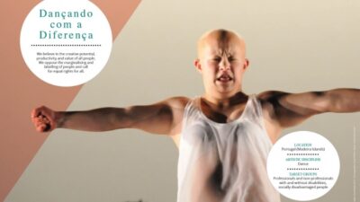 Man with arms outstretched and a white vest on, he has a shaved head.