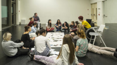 A group of people in a studio space sit on the floor looking at paper and print outs