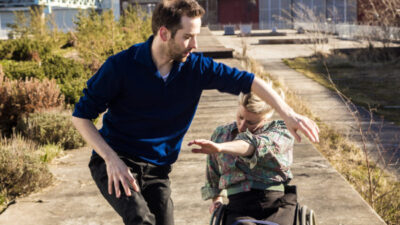 A female dancer with blonde hair using a wheelchair duets with a non-disabled male dancer outside in the sun.