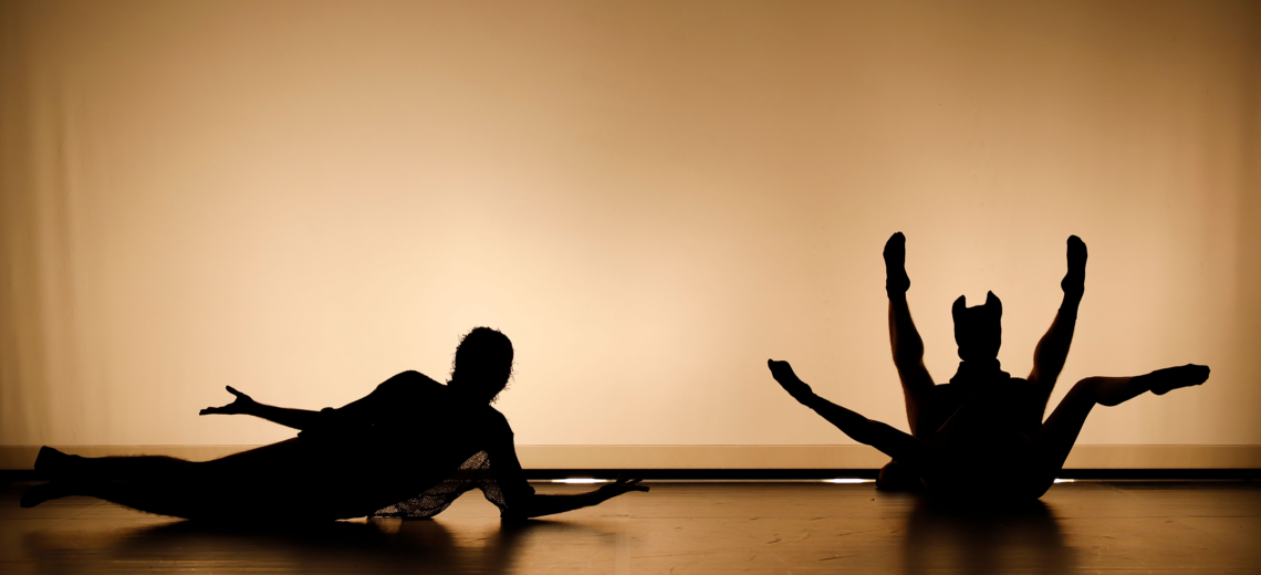 three performers are on a dark stage with an illuminated backdrop. The light is warm, in shades of yellow and orange and creates silhouettes with the performers' bodies