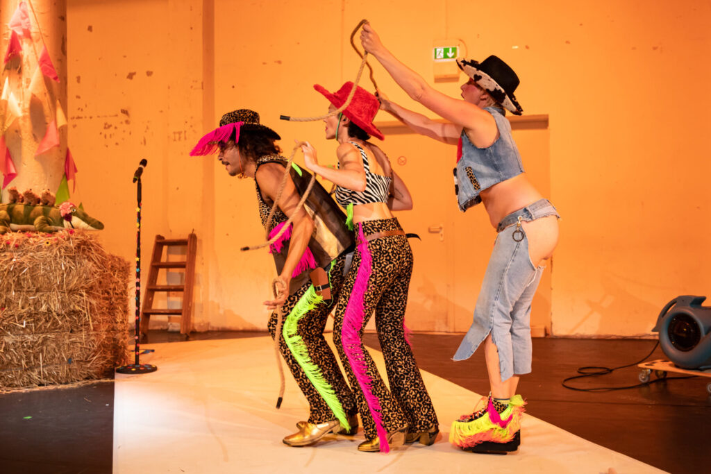 Three performers in colorful, eccentric cowboy-inspired costumes are lined up on stage, playfully pulling on ropes connected to each other. One wears leopard print with neon green and pink fringe, another wears a red fuzzy cowboy hat and zebra print top, while the third wears a denim outfit with exposed buttocks and bright platform shoes with fringe. The stage is decorated with hay bales, plush toys, and pink bunting, evoking a surreal rodeo atmosphere. The show: The Butching Cowboys by Anajara Amarante