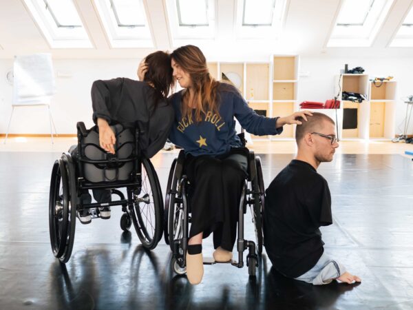 Two female dancers are on wheelchairs, one of them is holding the head of the other. The male artist, without arms and legs, is sitting on the floor next to one of the wheelchair. The dancer is gently touching his hair. They are in a dance studio and the overall mood is one of support