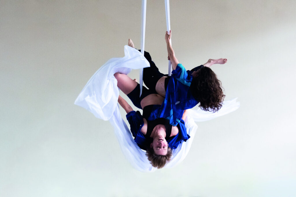Two aerial performers are suspended upside down, intertwined in midair using white aerial silks. Both wear flowing blue garments over black outfits. One performer has short hair and faces the camera, while the other, with long curly hair, arches back gracefully. The background is a soft, neutral tone, emphasizing the movement and elegance of the pose. Performance: With Or Without You by Fia Neises