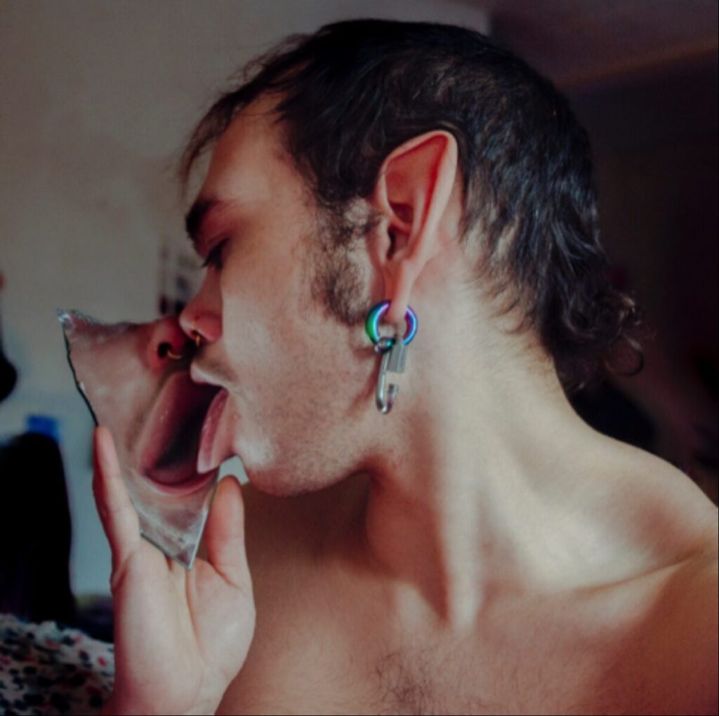 A shirtless person with facial hair and colorful hoop earrings is holding and licking a jagged shard of mirror. Their tongue and reflection appear distorted in the glass. The background is softly lit and out of focus. The show: FOR ALL INTENTS AND PURPOSES // BARE WITNESS // BOTTOMS UP by Rayne J. Raney