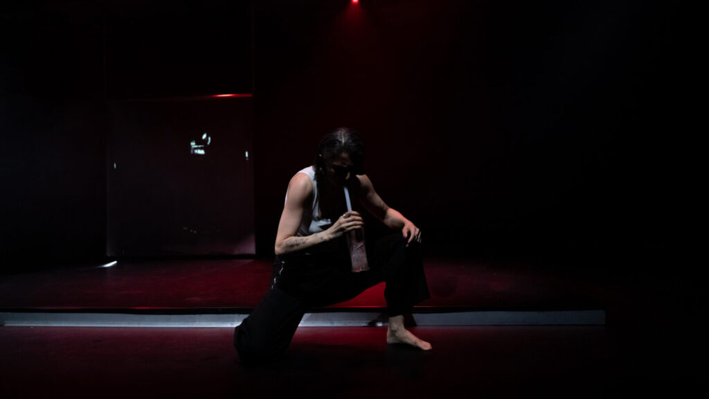 A solo performer on a dimly lit stage with dramatic red lighting. The person, wearing a white sleeveless top and dark pants, is captured mid-movement in a crouched or lunging position with one hand on the ground. The stage appears to have red steps or platforms, and the lighting creates a moody, theatrical atmosphere. Show: Rita Mazza The Voice