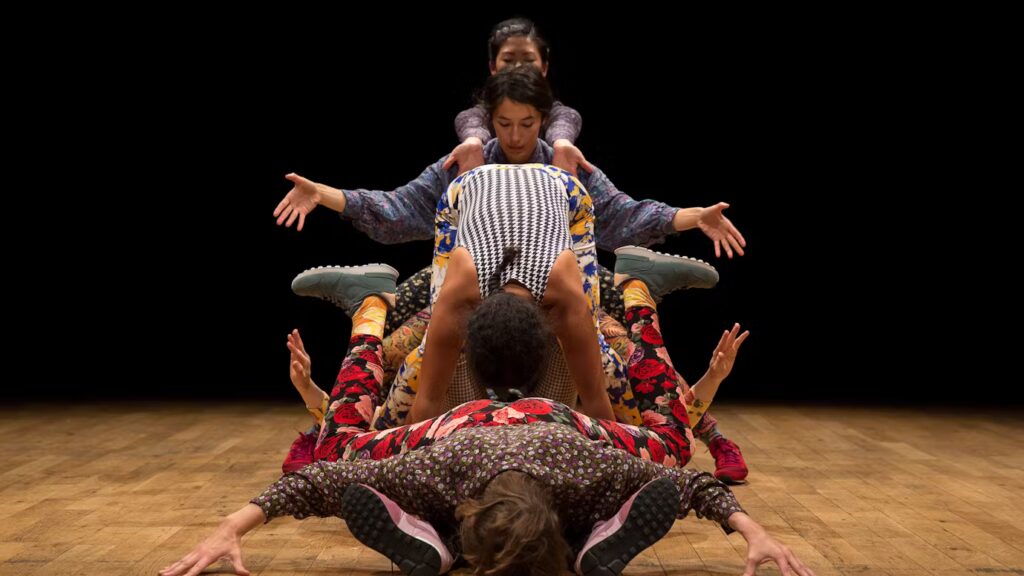 Four performers create a dynamic human tower on a wooden stage floor against a black background. At the base, a person in floral-patterned clothing lies face down with arms extended. Above them, a person in a black and white houndstooth top is positioned horizontally. In the middle, another performer in colorful patterned clothing extends their arms outward for balance. At the top, a fourth performer in light-colored clothing completes the formation with arms also outstretched, creating a sculptural arrangement of interconnected bodies in what appears to be a contemporary dance or physical theater performance. The show:Screensaver Series by J Neve Harrington
