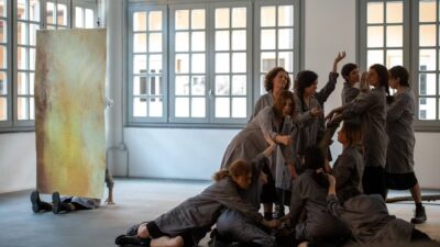 A group of women wearing grey coats and combat boots stand still in a bright industrial space with large windows. Their postures are rigid and solemn, evoking a sense of ritual. The atmosphere is stark and intense, aligning with the themes of resistance and female strength from the performance "Dies Irae – Concerto for Women and Hammers" by Gloria Dorliguzzo.