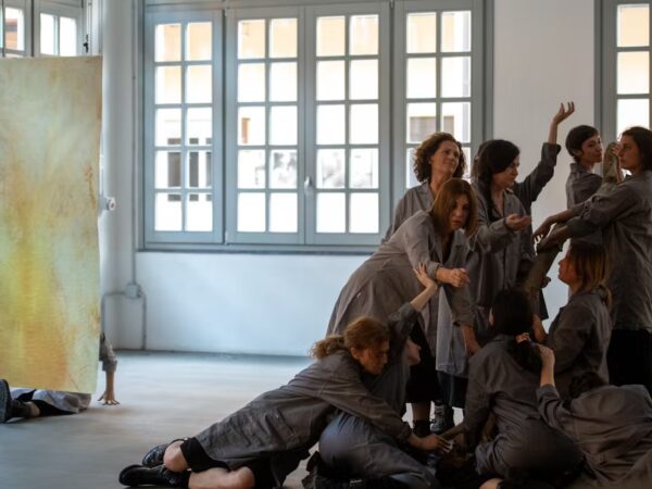A group of women wearing grey coats and combat boots stand still in a bright industrial space with large windows. Their postures are rigid and solemn, evoking a sense of ritual. The atmosphere is stark and intense, aligning with the themes of resistance and female strength from the performance "Dies Irae – Concerto for Women and Hammers" by Gloria Dorliguzzo.