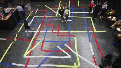 People walking and gathering around a floor installation made of intersecting red, blue, yellow and white tape in a black box space