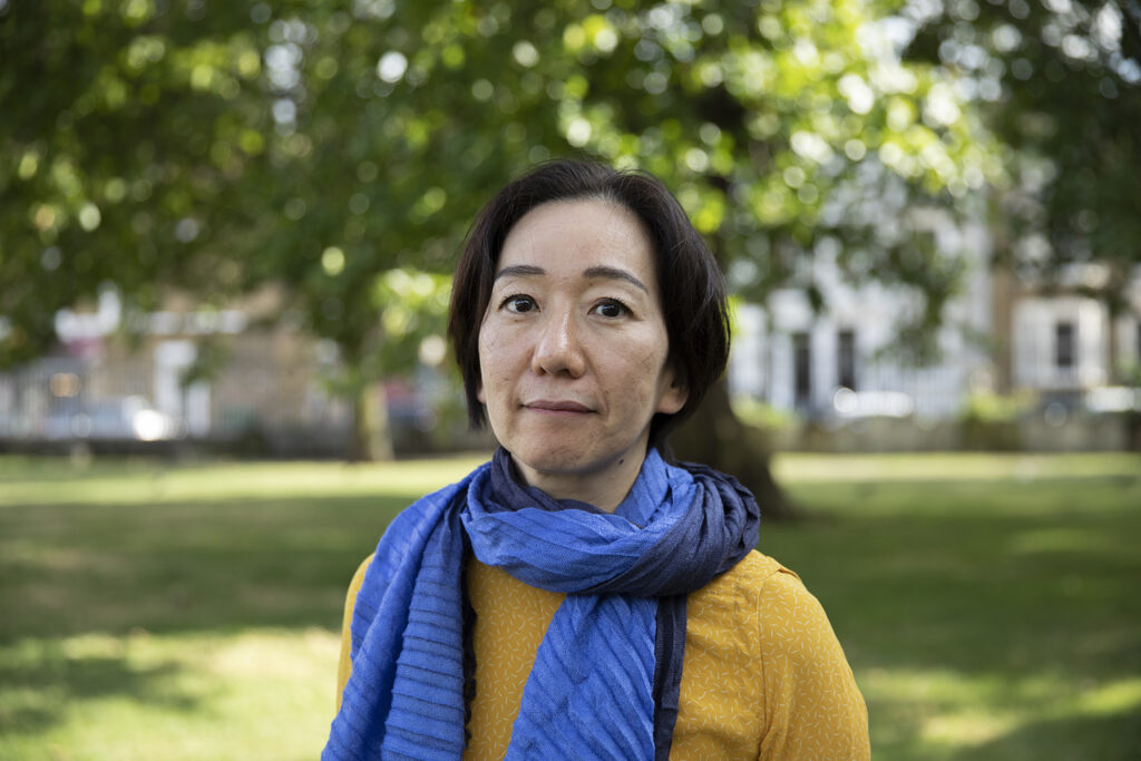 Chisato Minamimura wears a yellow jumper and a blue scarf. There is a park in the background