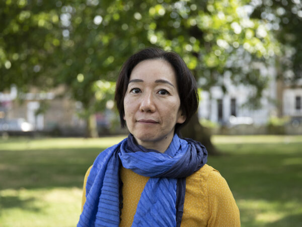 Chisato Minamimura wears a yellow jumper and a blue scarf. There is a park in the background