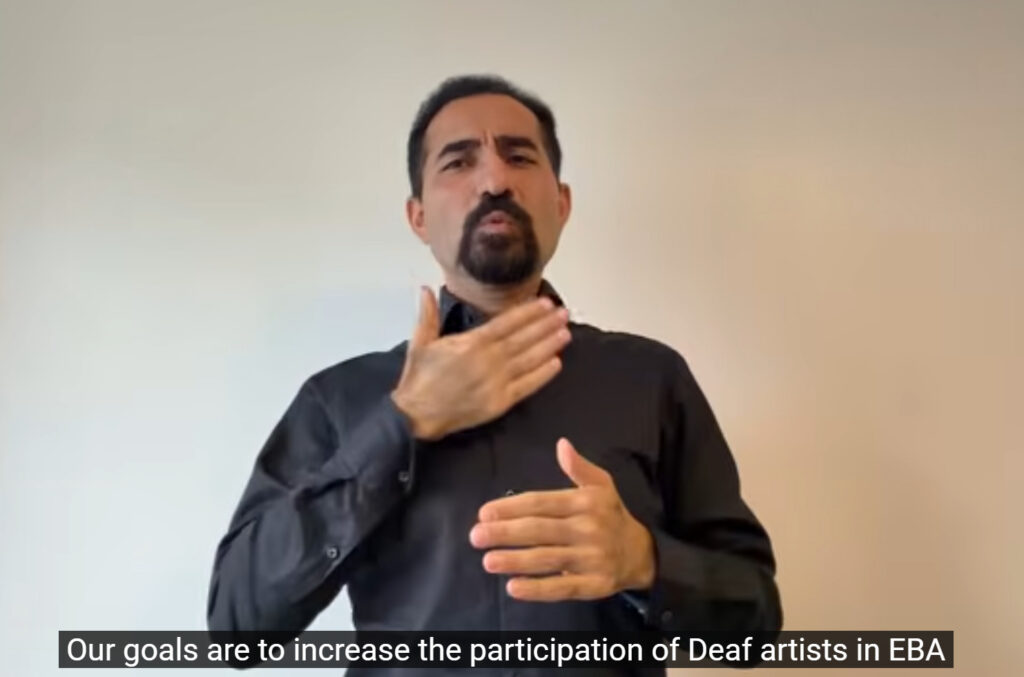 A Deaf man signing. He wears a black shirt. The background is white.