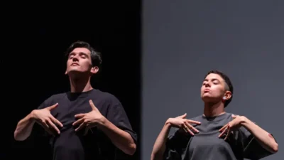 Two performers stand side by side on a stage, each wearing a loose dark-grey T-shirt. Both hold their hands near their chest with matching gestures, creating a sense of symmetry and connection. The background is divided between a dark black area and a light grey panel, highlighting the performers’ poses and the contrast of the stage design.