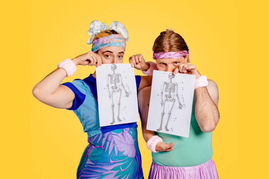 Two performers pose against a solid yellow backdrop, dressed in brightly coloured exercise-inspired clothing with matching headbands and wristbands. Each holds a black-and-white anatomical drawing of a human skeleton in front of their body. One performer wears a blue outfit with purple floral patterns, while the other wears a mint green top and a light pink skirt. The contrast between the playful costumes and the medical illustrations creates a striking visual reference to bodies, movement and representation.