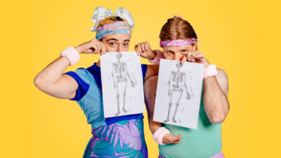 Two performers pose against a solid yellow backdrop, dressed in brightly coloured exercise-inspired clothing with matching headbands and wristbands. Each holds a black-and-white anatomical drawing of a human skeleton in front of their body. One performer wears a blue outfit with purple floral patterns, while the other wears a mint green top and a light pink skirt. The contrast between the playful costumes and the medical illustrations creates a striking visual reference to bodies, movement and representation.